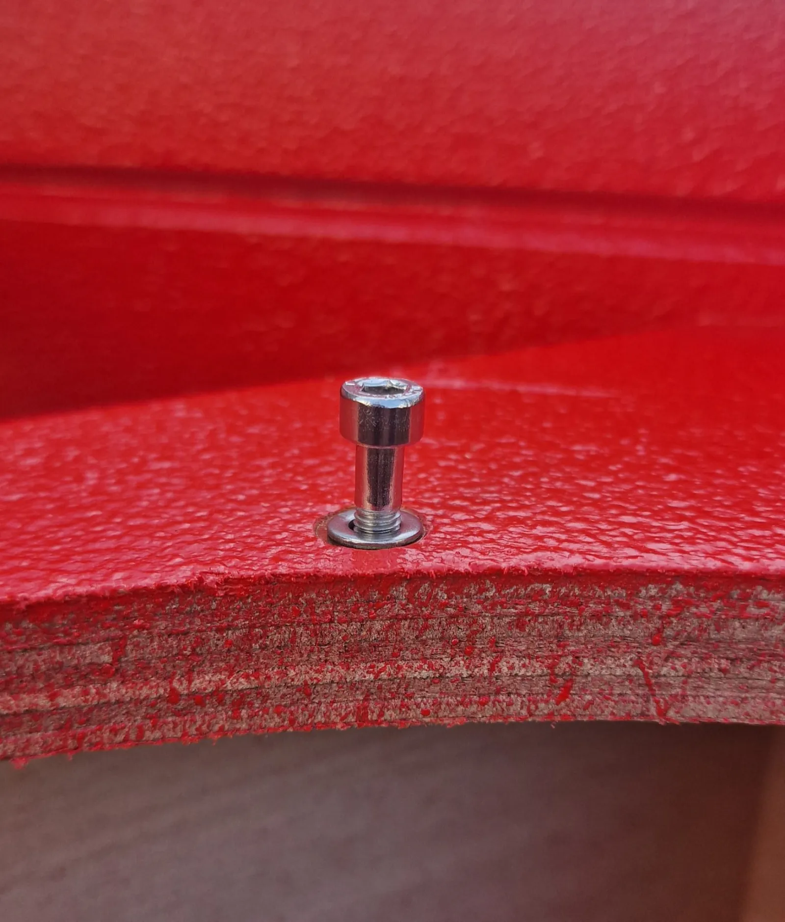 Close-up of a red textured speaker cabinet corner showing a stainless steel socket head bolt through the marine plywood edge